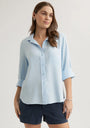 A woman with long brown hair wears a light blue Bella Dahl Lyocell Hemdbluse in an oversized cut and dark navy shorts. With her hands in her pockets, she looks slightly to the side and smiles softly against a plain, light background.