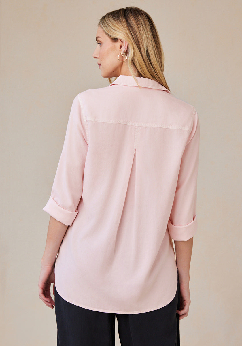 A woman with long blonde hair stands facing away, wearing a pale pink Bella Dahl Lyocell Hemdbluse with rolled-up sleeves, pleat in the back, oversized Schnitt, and relaxed fit, paired with black pants against a beige backdrop.