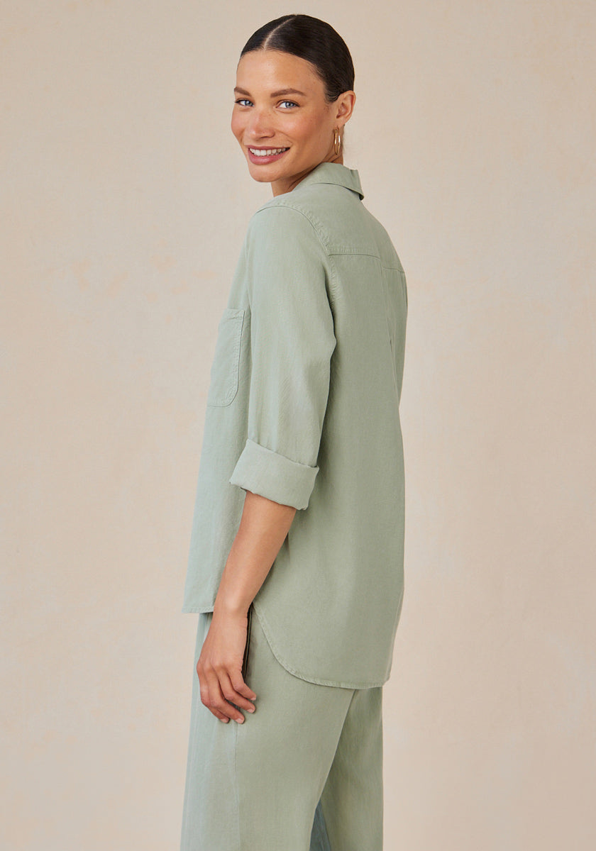A woman with medium skin and dark hair tied back smiles over her shoulder. She wears a light sage green Lyocell Hemdbluse with button placket and rolled sleeves by Bella Dahl, paired with matching wide-leg pants against a neutral beige background.