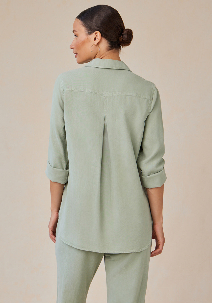A woman with dark hair in a low bun faces away, wearing a light sage green Bella Dahl Lyocell Hemdbluse with rolled-up sleeves and matching pants. The shirt has a back pleat, relaxed fit, subtle Knopfleiste, set against a light beige background.