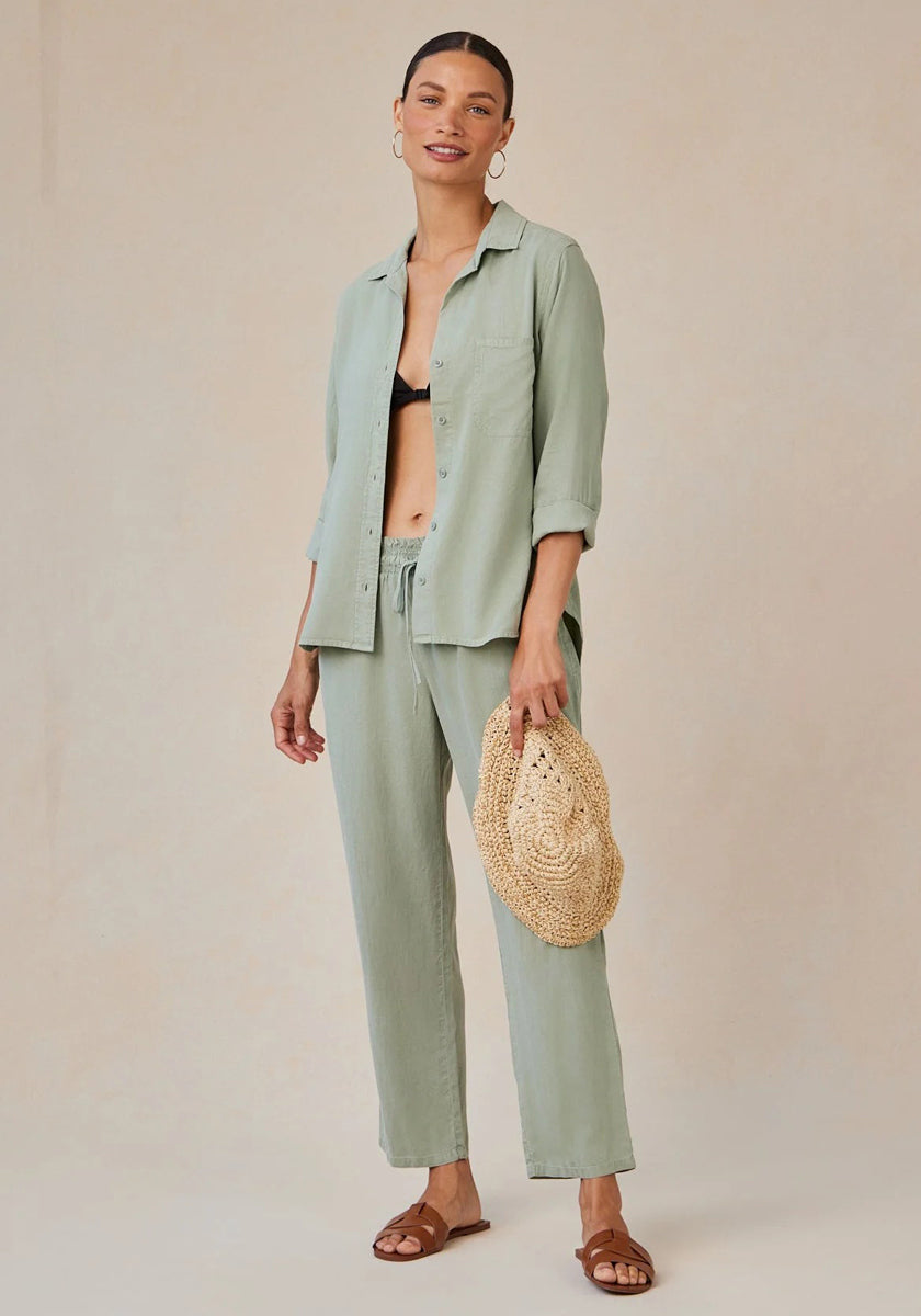 A woman smiles in a relaxed light sage green Bella Dahl Lyocell Hemdbluse with Knopfleiste over a black bralette, paired with matching drawstring pants, brown sandals, large hoop earrings, and a woven straw bag. Her hair is pulled back against a plain beige backdrop.