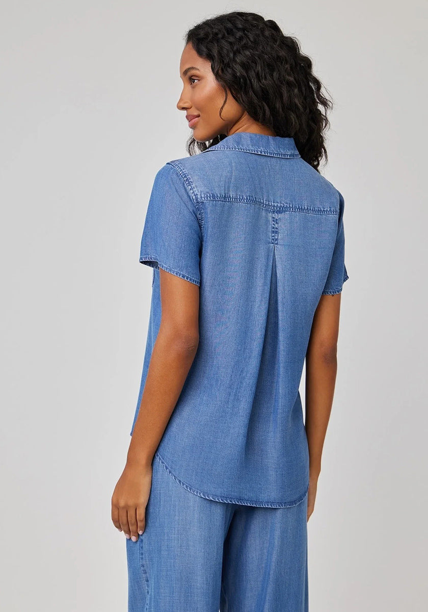A woman with long, curly dark hair stands facing away, wearing a light blue Bella Dahl Lyocell Kurzarmbluse with Hemdkragen and matching loose pants. The blouse features a back pleat and seam detailing, set against a plain light gray background.