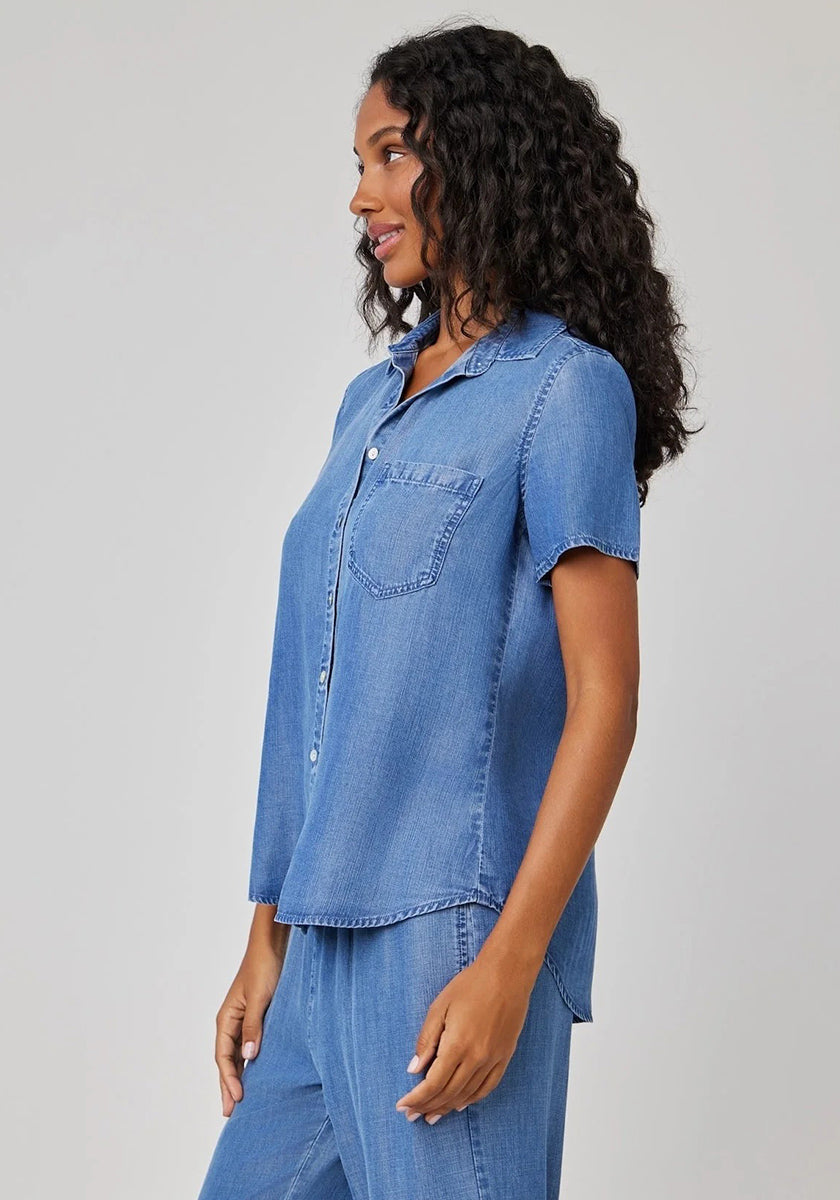 A woman with long curly hair models a Bella Dahl Kurzarmbluse aus Lyocell with Hemdkragen and chest pocket, paired with blue denim pants. She stands sideways, smiling against a plain light gray background.