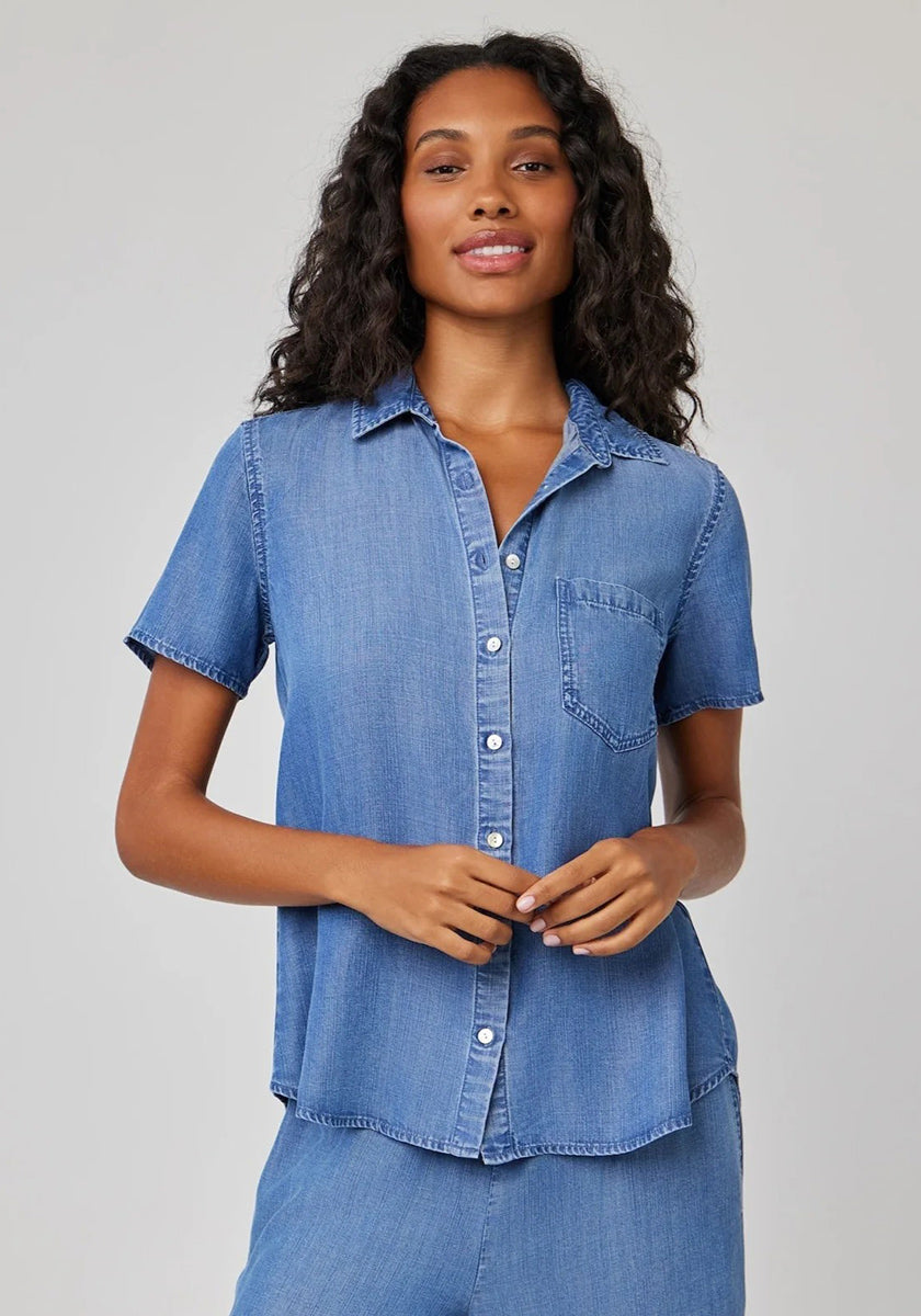A woman with long, wavy hair wears a Bella Dahl short-sleeve lyocell blouse with shirt collar and chest pocket in light blue denim. She stands against a plain light gray background, smiling slightly with her hands at her waist.