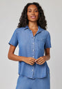 A woman with long, wavy hair wears a Bella Dahl short-sleeve lyocell blouse with shirt collar and chest pocket in light blue denim. She stands against a plain light gray background, smiling slightly with her hands at her waist.