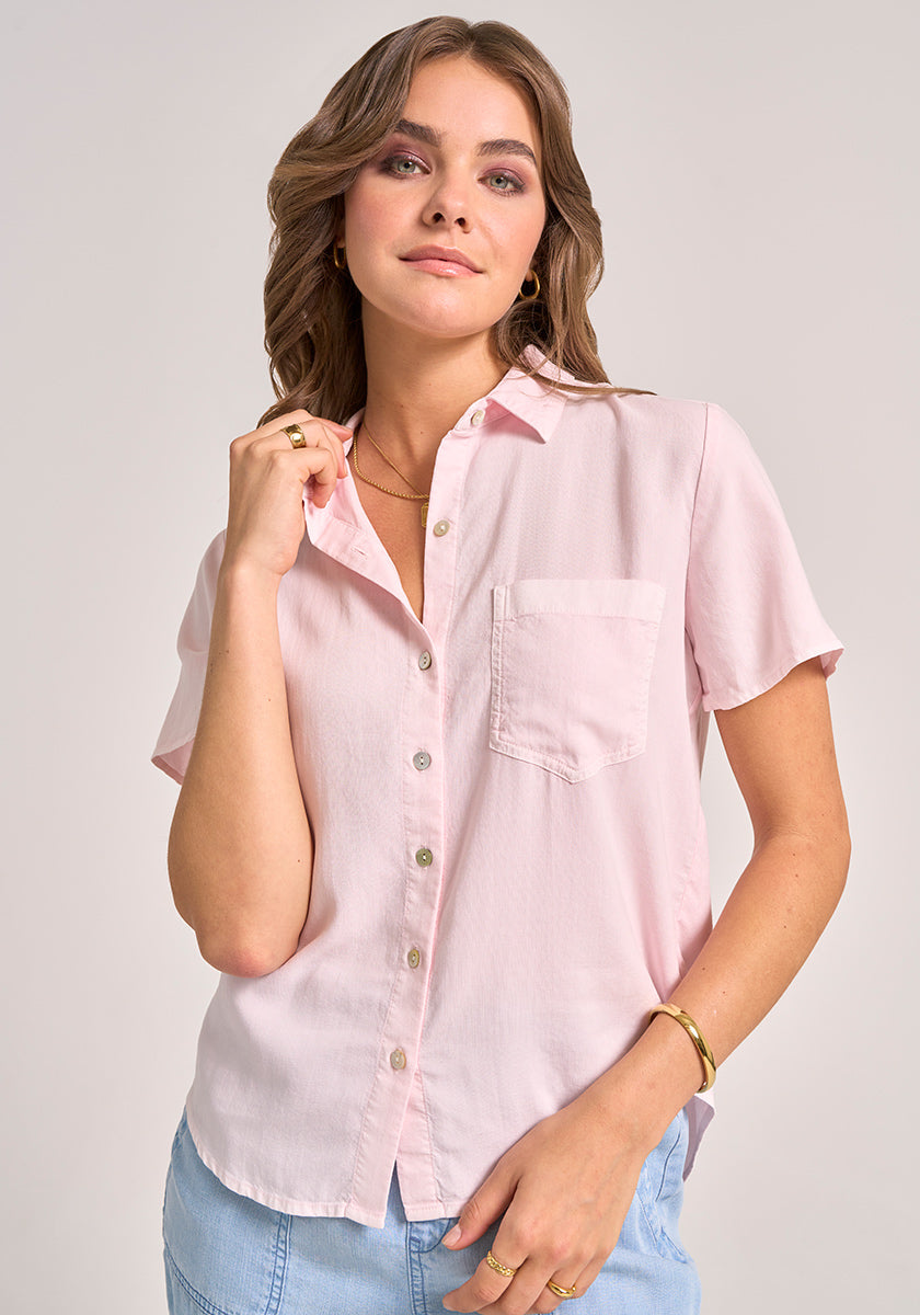 A woman with wavy brown hair wears a pale pink Bella Dahl Kurzarmbluse aus Lyocell mit Knopfleiste und Brusttasche. She poses relaxed, holding her collar, accessorized with gold jewelry against a light gray background.