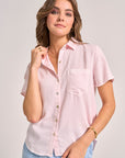 A woman with wavy brown hair wears a pale pink Bella Dahl Kurzarmbluse aus Lyocell mit Knopfleiste und Brusttasche. She poses relaxed, holding her collar, accessorized with gold jewelry against a light gray background.