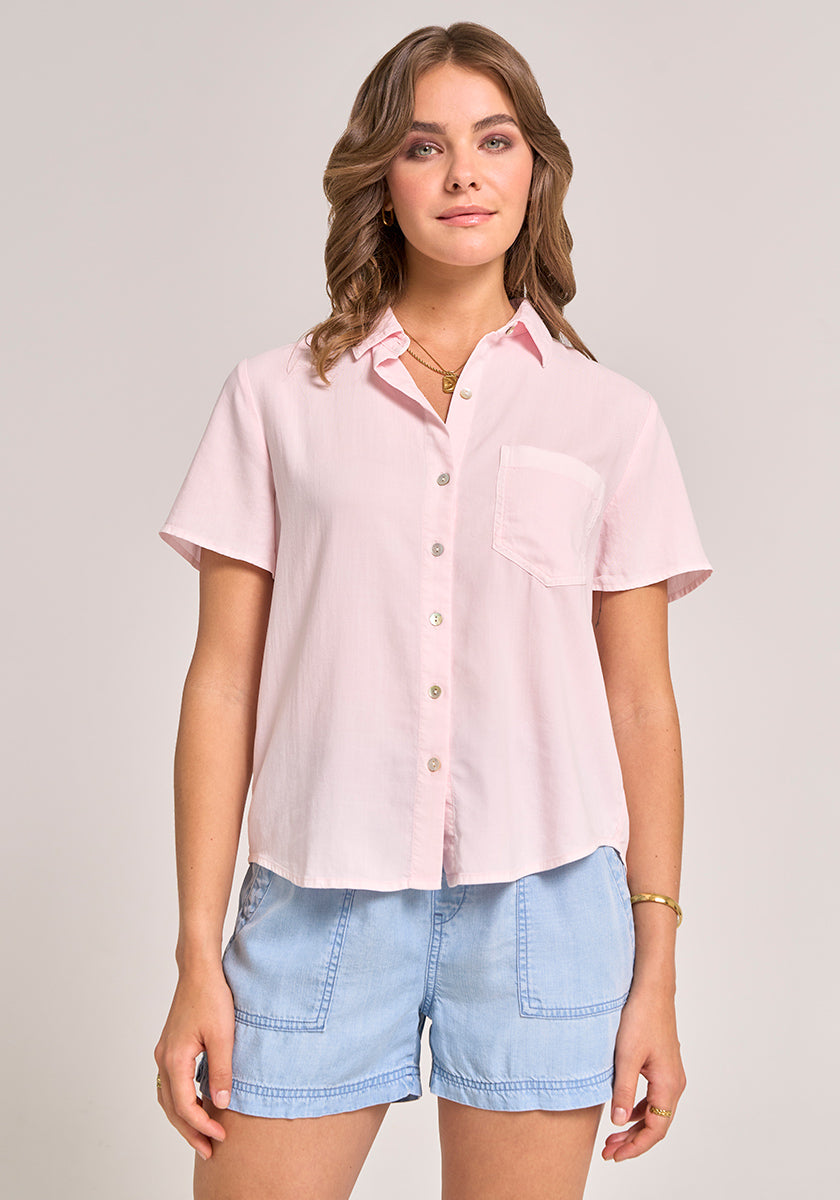 A woman with wavy brown hair and light makeup faces forward in a Bella Dahl light pink Lyocell Kurzarmbluse with Hemdkragen and left chest pocket, paired with light blue denim shorts. She accessorizes with a bracelet and necklace.