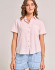 A woman with wavy brown hair and light makeup faces forward in a Bella Dahl light pink Lyocell Kurzarmbluse with Hemdkragen and left chest pocket, paired with light blue denim shorts. She accessorizes with a bracelet and necklace.
