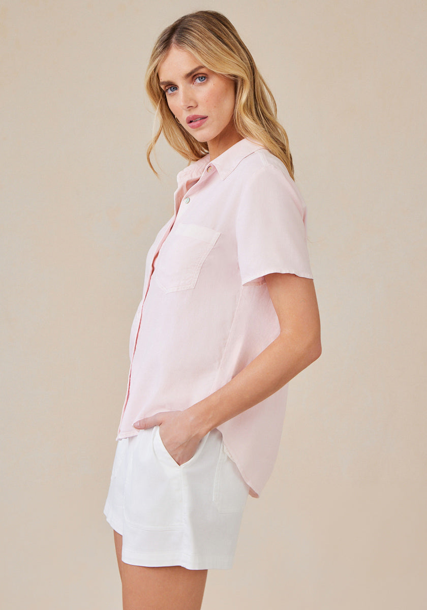 A woman with long blonde hair stands sideways, looking at the camera. She wears a light pink Bella Dahl Lyocell blouse with hemdkragen and chest pocket, paired with white shorts. Her left hand is in her pocket; the background is plain beige.