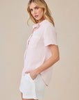 A woman with long blonde hair stands sideways, looking at the camera. She wears a light pink Bella Dahl Lyocell blouse with hemdkragen and chest pocket, paired with white shorts. Her left hand is in her pocket; the background is plain beige.