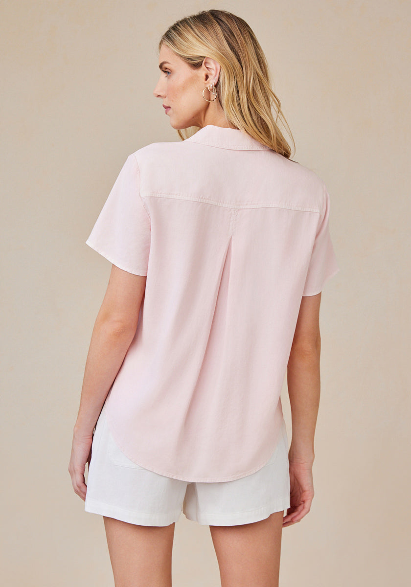 A woman with long blonde hair faces away, wearing a pale pink Bella Dahl Lyocell blouse with short sleeves, shirt collar, back pleat, and curved hem, paired with white shorts against a light beige background.