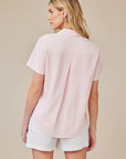 A woman with long blonde hair faces away, wearing a pale pink Bella Dahl Lyocell blouse with short sleeves, shirt collar, back pleat, and curved hem, paired with white shorts against a light beige background.