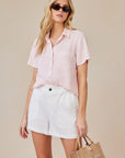 A woman with long blonde hair wears sunglasses, a light pink Lyocell blouse with a shirt collar by Bella Dahl, and high-waisted white shorts. She holds a small brown woven handbag. The background is neutral beige.