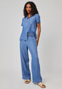 A smiling woman with curly hair wears a Bella Dahl short-sleeve, button-up denim shirt with a chest pocket, Lyocel-Hose in Denimoptik, and beige slide sandals. She stands against a light background with one hand in her pocket.