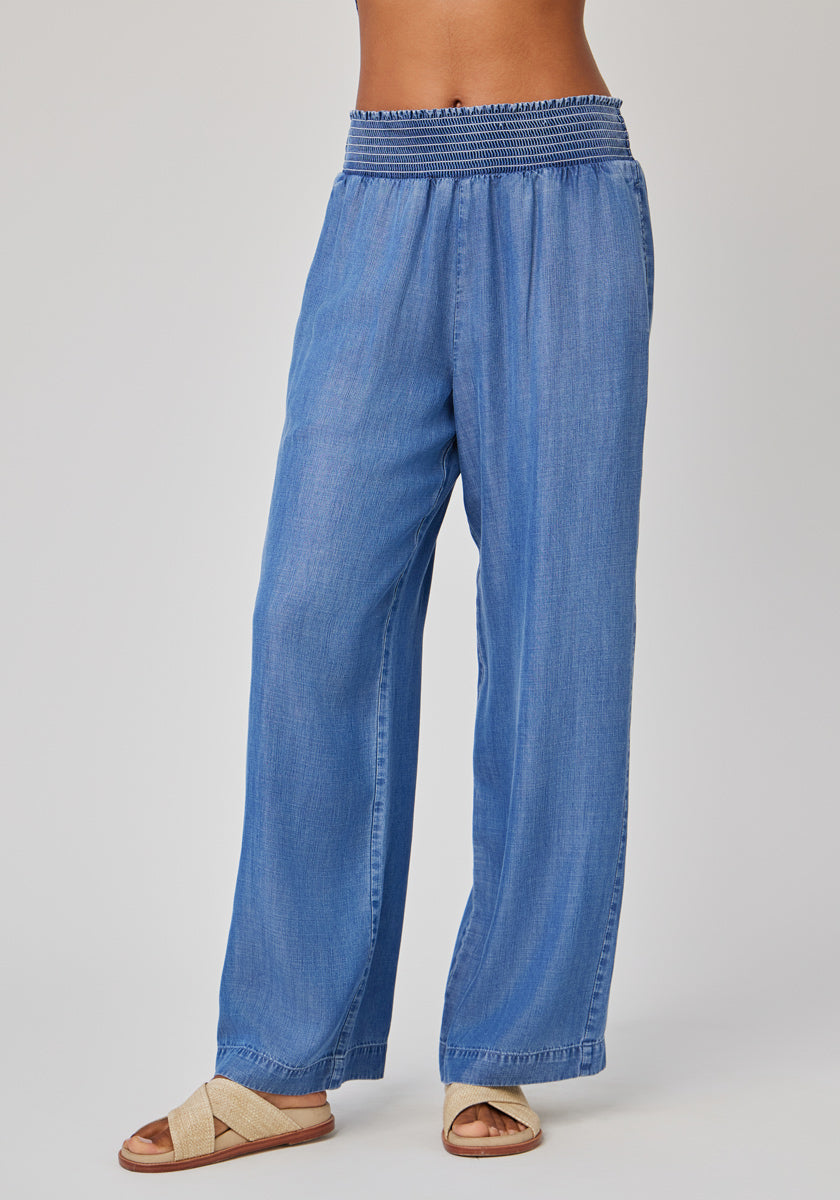 A person models Bella Dahl’s loose, light blue Lyocel-Hose in Denimoptik with a smocked elastic waistband and wide legs, paired with beige crisscross sandals. Torso and head are not visible; background is plain and light-colored.