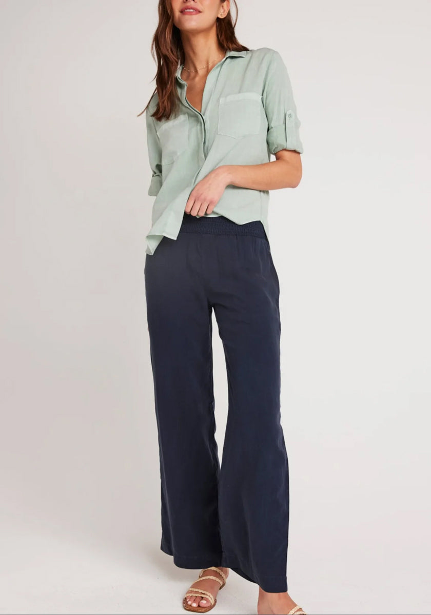A woman stands against a plain light background wearing a Bella Dahl light green button-up shirt with rolled sleeves, navy wide-leg denim-look pants, and tan flat sandals. Her hair is loose and wavy, with her face partially out of frame.
