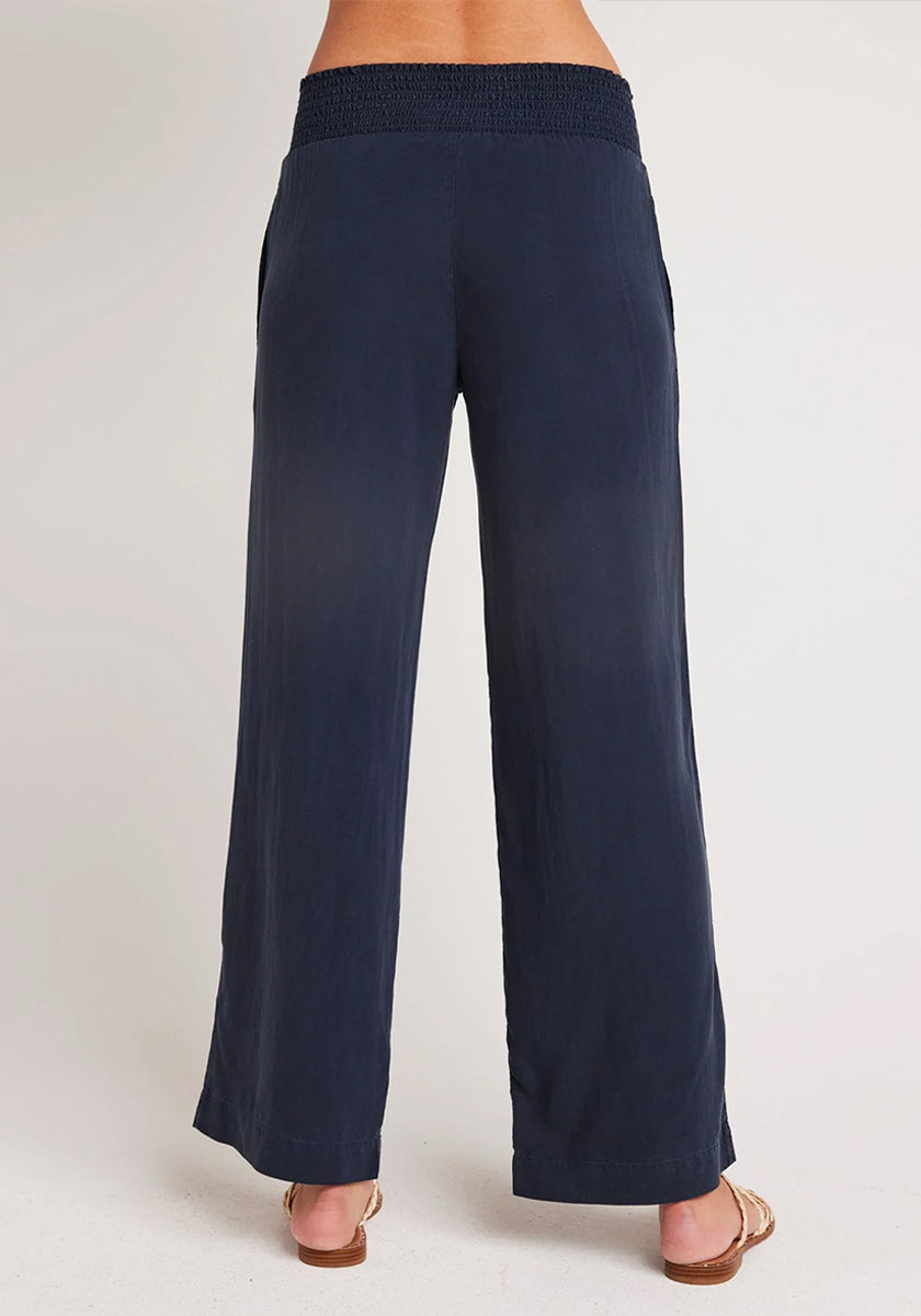 A person stands barefoot on a light background, facing away from the camera, wearing Bella Dahl’s navy blue wide-leg Lyocell pants in denim look with a high smocked waistband and relaxed fit.