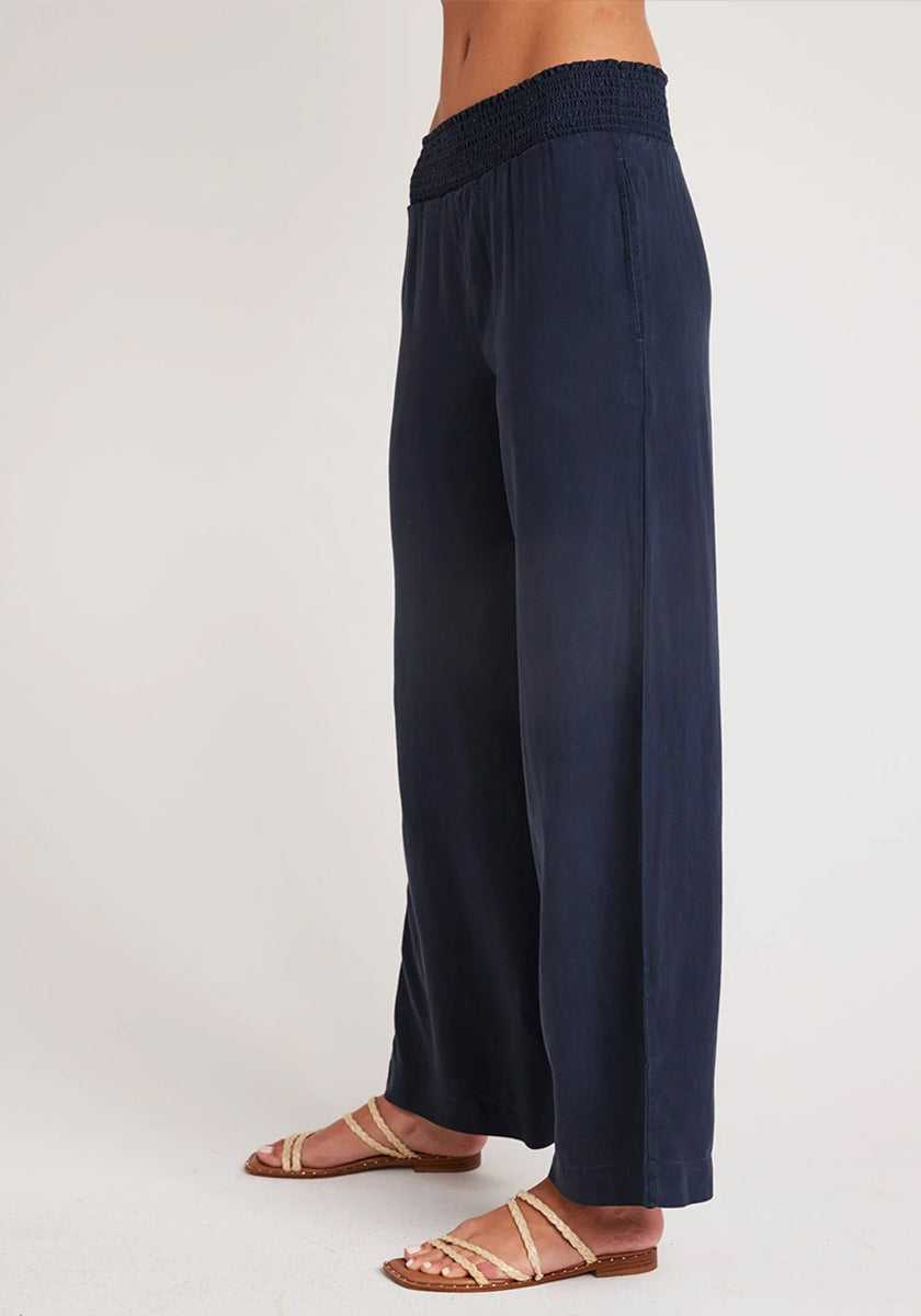 A person in profile wears Bella Dahl’s navy blue wide-leg pants in denim look, featuring a smocked waistband. Their feet are in flat tan sandals with gold crisscross straps. The plain light background highlights only their lower torso and legs.
