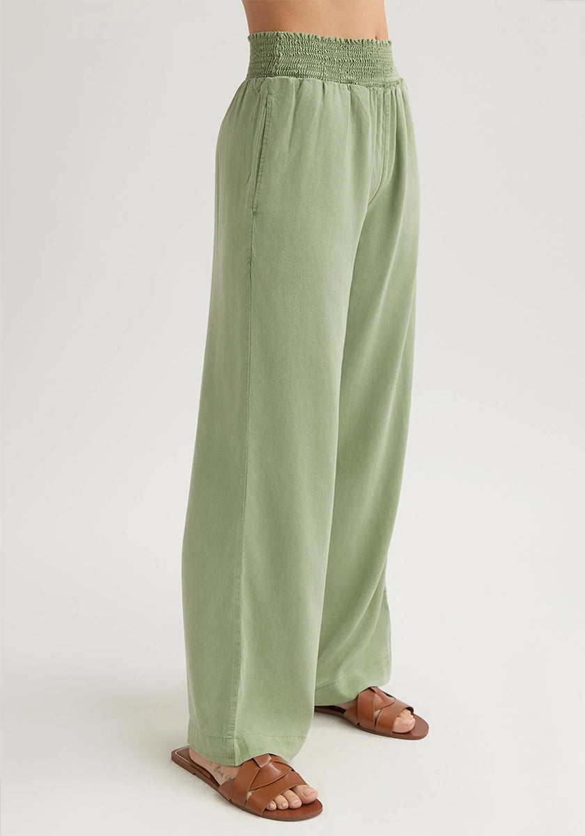 A person stands on a white background, wearing Bella Dahl’s loose, light green Lyocell denim-look pants with an elastic waistband and side pockets. Brown flat sandals complete the look. Only the lower torso and legs are visible.