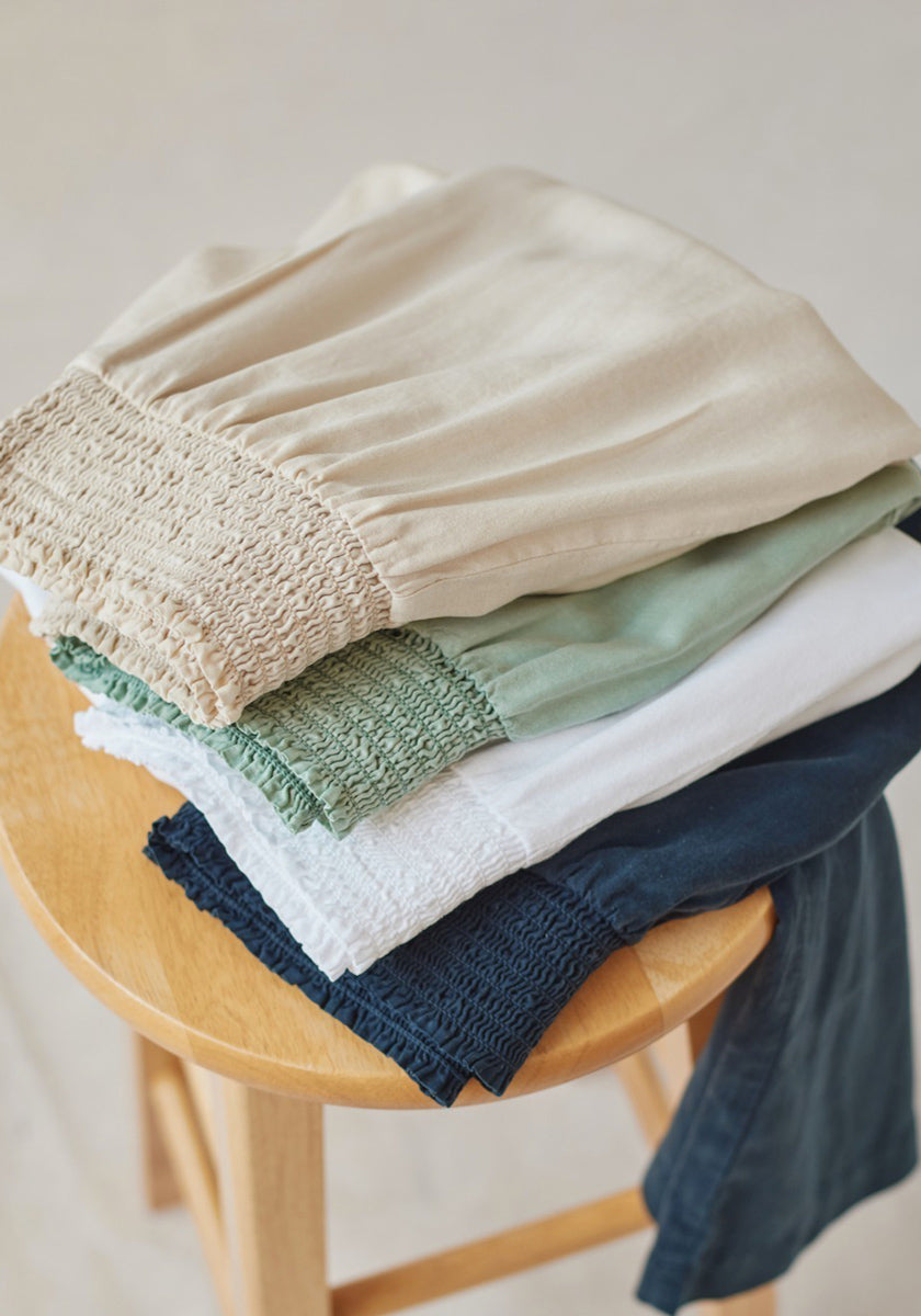 A neatly folded stack of five Bella Dahl weite Hosen in beige, light green, white, light blue, and navy sits on a wooden stool, showing smocked waistbands—a soft display of Lyocell and Denimoptik styles.