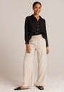 A woman smiles against a light beige background, wearing a black long-sleeve button-up Bella Dahl shirt, beige wide-leg pants with a shirred waistband, and black slide sandals. Her hair is pulled back, hands resting naturally at her sides.