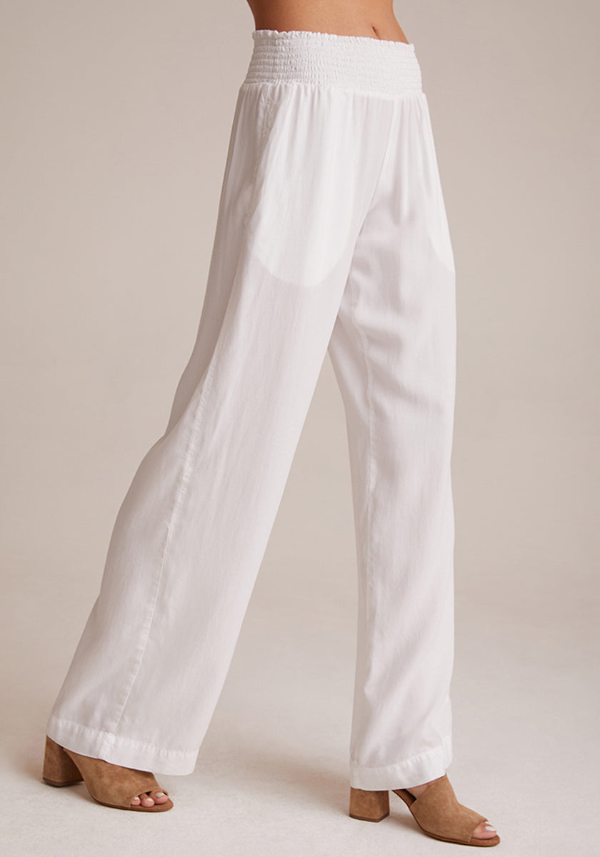 A person stands against a light background wearing loose-fitting, white, wide-leg Bella Dahl Lyocell pants with a smocked elastic waistband. The slightly sheer pants are paired with tan, open-toed block-heeled sandals. Upper body is not visible.