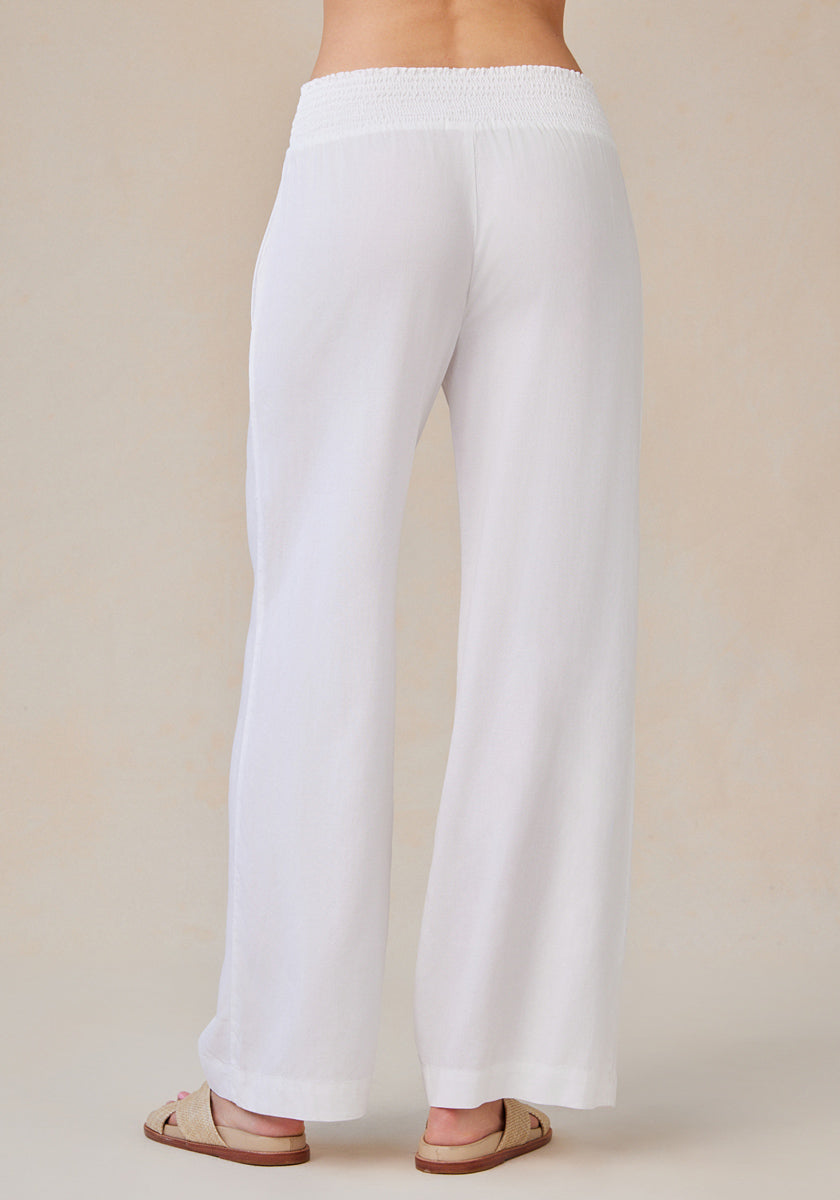 A person faces away, displaying the back of loose-fitting white Bella Dahl Lyocell wide-leg pants with a broad smocked waistband in denim look. They wear tan slide sandals against a light beige neutral backdrop; both floor and pants are light-colored.