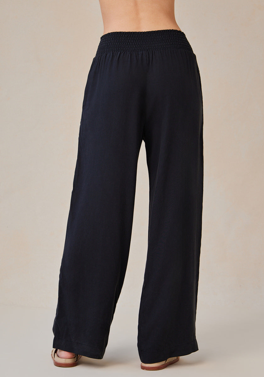 A person stands facing away, wearing loose-fitting, high-waisted black Bella Dahl Lyocell pants with a wide smocked waistband. The relaxed fit trousers reach the tops of their sandals against a plain, light-colored background.