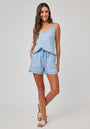 A woman with long wavy brown hair smiles with one hand in her pocket, wearing a light blue Bella Dahl Lyocell camisole top with lace trim and matching drawstring shorts. She pairs them with hoop earrings and beige slide sandals against a plain gray background.