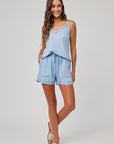 A woman with long wavy brown hair smiles with one hand in her pocket, wearing a light blue Bella Dahl Lyocell camisole top with lace trim and matching drawstring shorts. She pairs them with hoop earrings and beige slide sandals against a plain gray background.