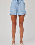 A person with medium brown skin faces forward, wearing Bella Dahl light blue Lyocell denim shorts with an elastic waistband and large front pockets, plus beige slip-on sandals. The background is plain white.