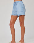A person stands barefoot, facing sideways, wearing Bella Dahl light blue denim drawstring shorts with a relaxed fit and large front pockets, paired with cream woven slide sandals. The background is plain white.