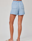 A person is shown from the waist down, facing away, wearing light blue Bella Dahl Lyocell denim shorts with a loose fit, two back pockets, and an elastic waistband. Light sandals complete the look against a plain white background.