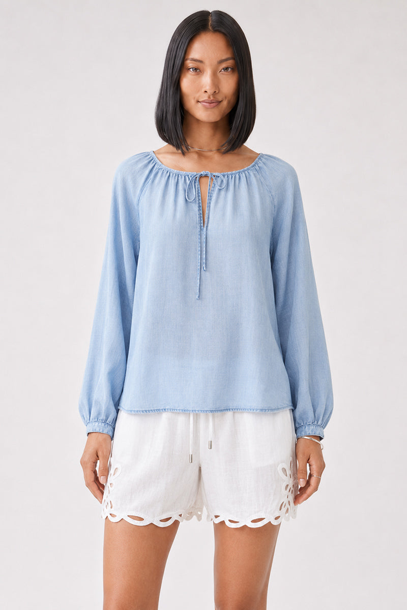 Eine Frau mit glattem schwarzem Haar trägt eine hellblaue Lyocell-Bluse von Bella Dahl mit Rundhalsausschnitt und langen Ärmeln, dazu weiße Shorts mit gewellten, bestickten Säumen. Sie steht mit dem Gesicht nach vorne vor einem schlichten, hellen Hintergrund und blickt mit neutralem Gesichtsausdruck in die Kamera.