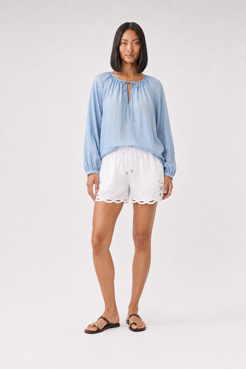 Eine Frau steht mit dem Blick nach vorne und trägt eine hellblaue Bella Dahl Lyocell-Bluse mit Rundhalsausschnitt und dezenten Ballonärmeln, dazu weiße Shorts mit Wellenrand und braune Sandalen. Sie hat glattes, schulterlanges schwarzes Haar und einen neutralen Gesichtsausdruck und steht vor einem hellgrauen Hintergrund.