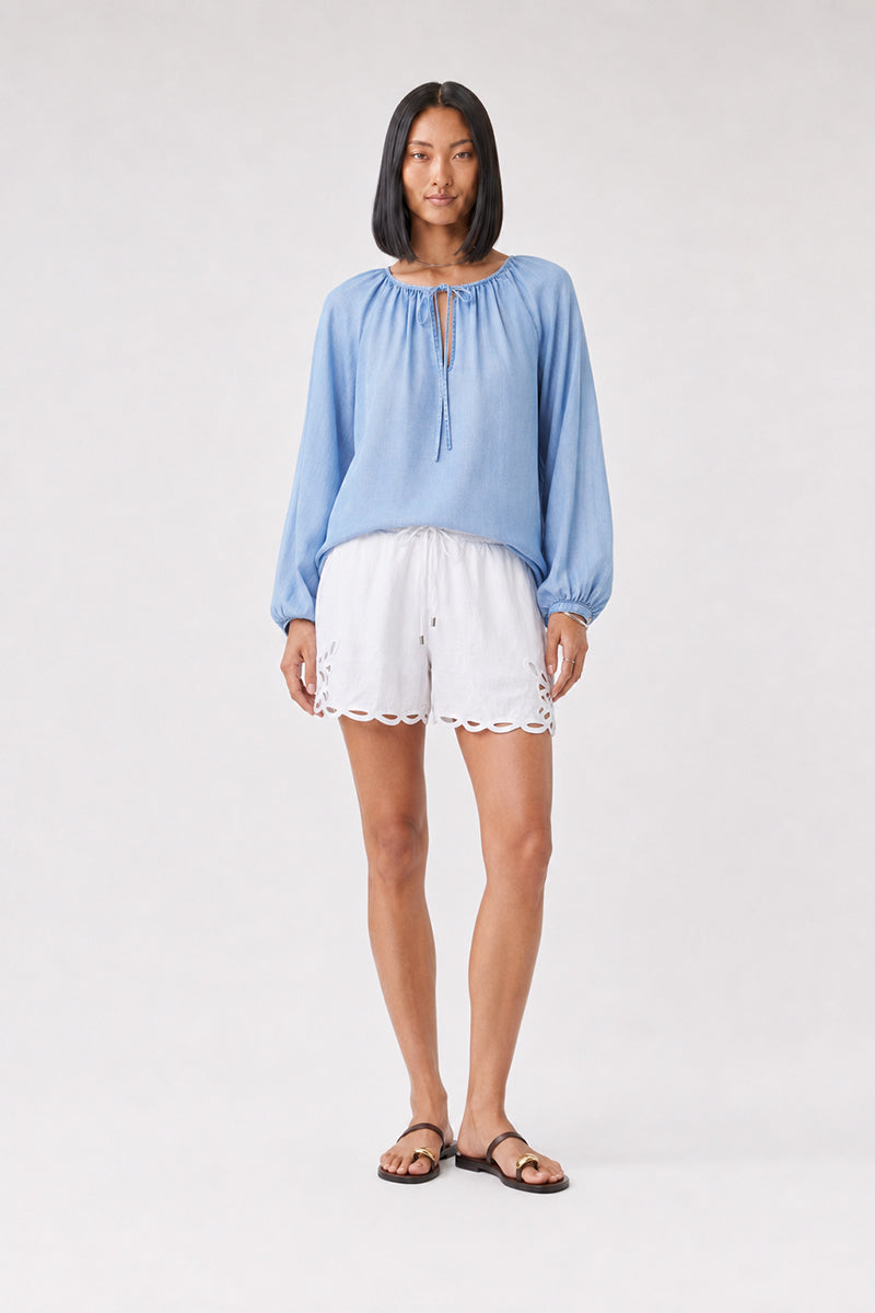 Eine Frau steht vor einem schlichten Hintergrund und trägt eine locker sitzende blaue Lyocell-Bluse von Bella Dahl mit Ballonärmeln und einem charmanten Rundhalsausschnitt, dazu weiße Shorts mit Wellenrand und braune Sandalen. Sie hat einen schulterlangen Bob und einen entspannten, neutralen Gesichtsausdruck, wobei ihre Arme an den Seiten herunterhängen.