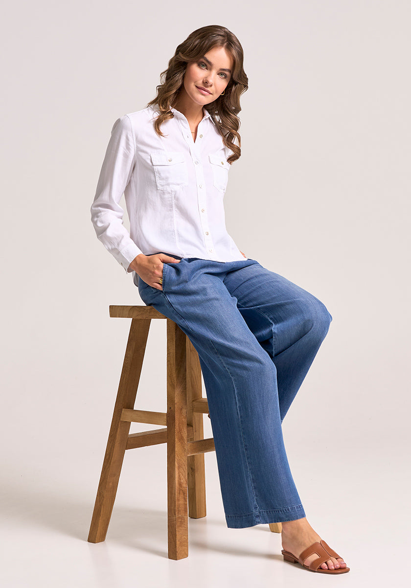 A woman with wavy brown hair sits on a wooden stool, wearing a white Lyocell Bluse in Jacken-Stil by Bella Dahl, wide-leg blue jeans, and brown sandals. She smiles slightly with her left hand in her pocket against a plain light gray background.