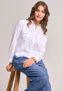 A woman with wavy brown hair sits on a wooden stool, wearing a white Bella Dahl Lyocell Bluse im Jacken-Stil with front pockets and loose blue jeans. She accessorizes with gold hoop earrings, a bracelet, and ring, gazing right with a relaxed expression.