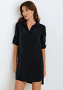 A woman with long brown hair wears a loose-fitting, black Lyocell V-neck dress with rolled-up sleeves from Bella Dahl, standing against a plain white background and gazing slightly left with a relaxed expression.