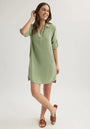 A woman stands against a plain light background, smiling in a sage green Bella Dahl Lyocell A-line shirt dress with rolled-up sleeves and a partial button placket. She touches her hair, legs crossed, wearing brown slide sandals.