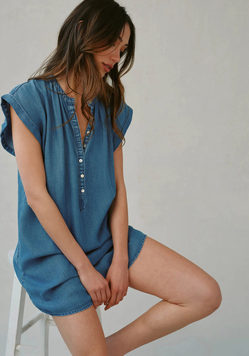 A woman with long brown hair sits on a white stool against a plain, light background, wearing a loose-fit blue denim dress by Bella Dahl with Denimoptim and Henley-Ausschnitt details, her head tilted down and hands gently in her lap.