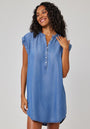 A woman with curly hair smiles in a Bella Dahl short dress, crafted from blue Denimoptim Lyocell with a buttoned placket. She stands against a plain light background, one hand touching her hair and the other by her side.