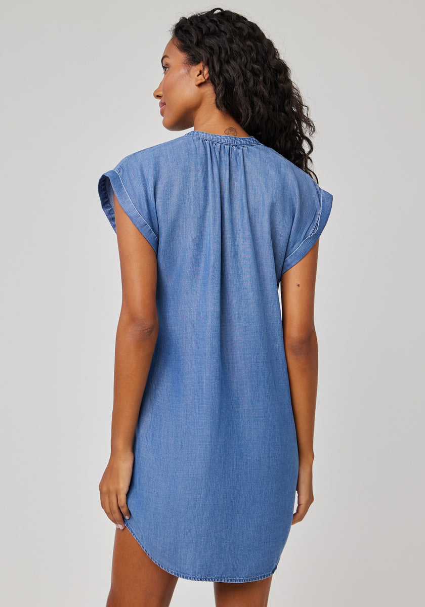 A woman with long wavy dark hair faces away from the camera, wearing a Bella Dahl short Lyocell-denim dress with a loose fit, gathered neckline, and short sleeves against a plain light gray background.