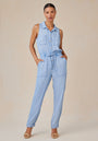 Against a beige background, a woman models the Bella Dahl sleeveless light blue denim-look jumpsuit with large front pockets, belted waist, and button-up front. She completes the look with tan heeled sandals and gold hoop earrings.