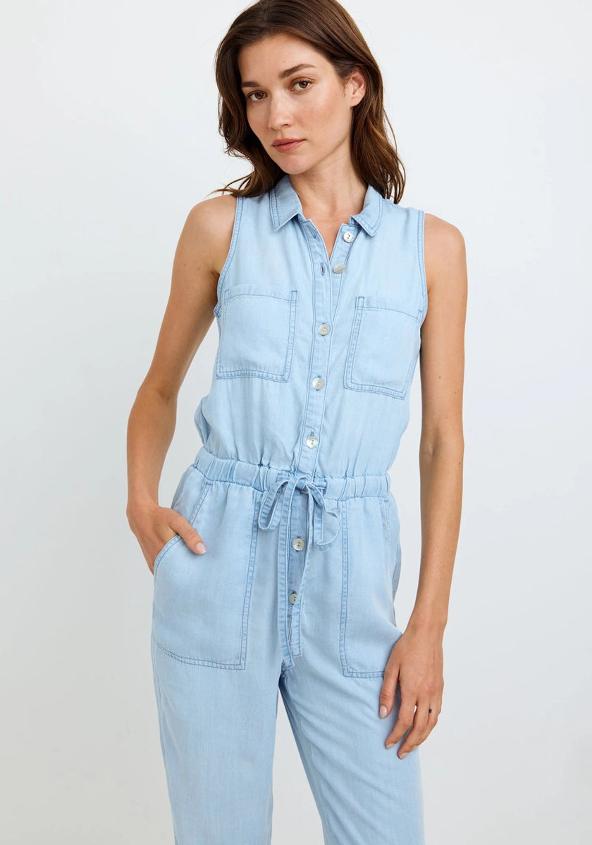 A woman poses against a white background wearing a Bella Dahl sleeveless light blue Lyocell jumpsuit in denim look with button-down front, two chest pockets, drawstring waist, and side pockets, her hand in one pocket and a neutral expression.