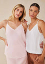 Two women smile at the camera in Bella Dahl camisoles with thin straps and frayed edges—one in pale pink with a skirt, the other in white layered over brown pants. The background is plain beige.