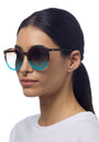 A woman with straight dark hair pulled back wears Okkias Berlino Sonnenbrille—round, oversized tortoiseshell frames with blue gradient UV 400 lenses. She wears a plain white top against a white background, facing slightly right.