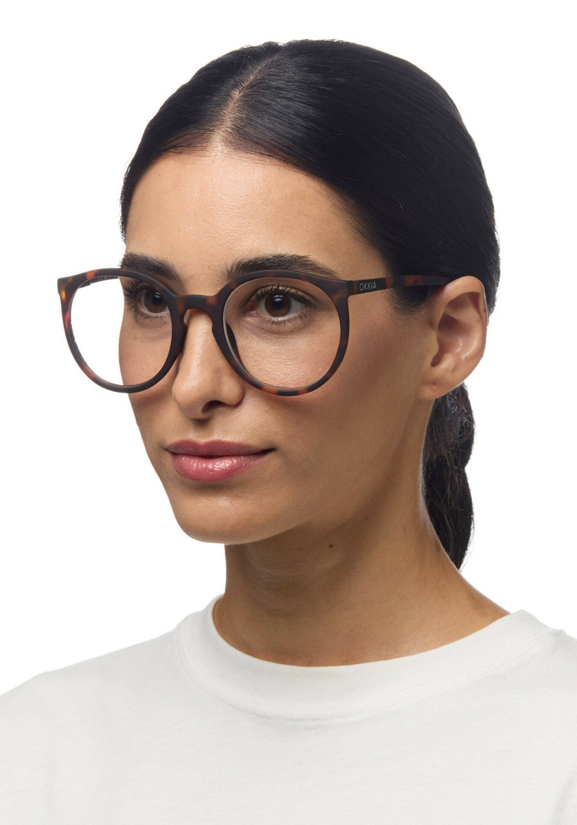 A woman with straight dark hair pulled back wears large, round Okkia reading glasses in a tortoiseshell pattern and a white top, facing slightly right with a relaxed expression against a plain white background.