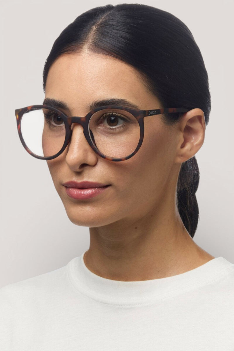 Eine Frau mit glattem, dunklem, nach hinten gebundenem Haar trägt eine Okkia Lesebrille mit einem runden, braunen Schildpattmuster und ein weißes Hemd. Sie blickt leicht nach rechts vor einem hellgrauen Hintergrund, ihr neutraler Look passt zur Soft-Touch-Oberfläche der Brille.